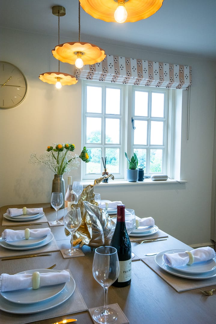 Kitchen table set for dinner with a vase with fresh flowers, plates, napkins, glasses and a bottle of red wine. There is a window with a pretty blind