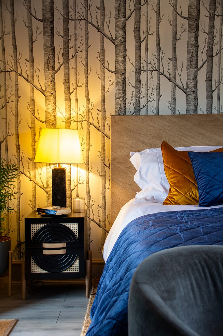 A bed dressed with white linen and a blue bed throw. Next to the bed is bedside table with a table lamp. Behind the bed is a wall with tree design wallpaper
