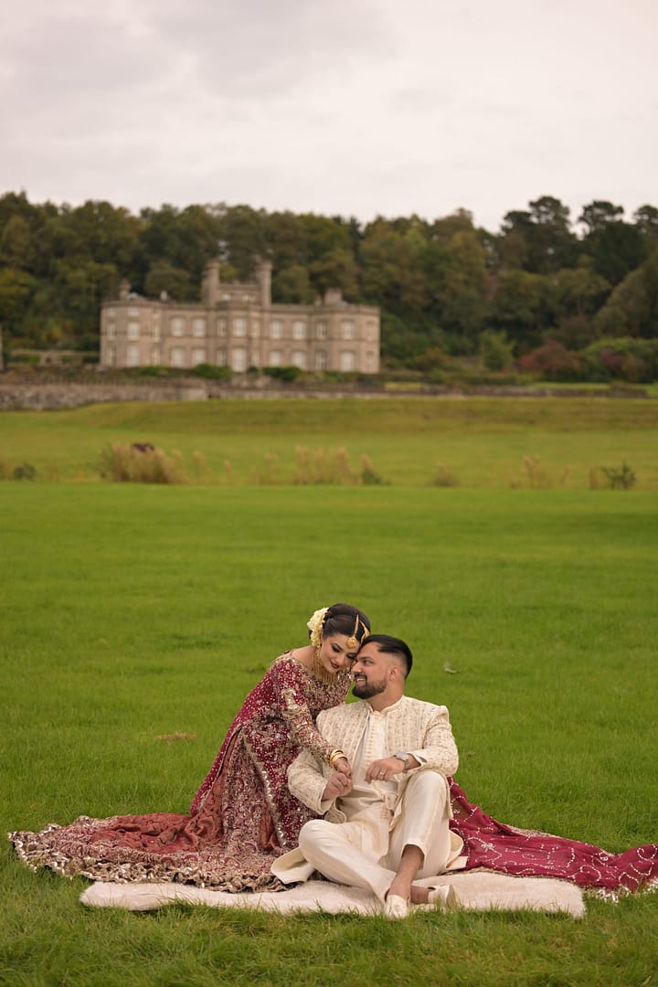 A beautiful South Asian wedding couple poses in an open green field with Bolesworth Castle in the background. The bride wears a stunning deep red and gold bridal lehenga with intricate embroidery, complemented by traditional jewelry and a matching veil, while the groom is dressed in a regal ivory sherwani with delicate gold embroidery. They sit on a plush white blanket, sharing an intimate and loving moment in this luxurious outdoor wedding shoot.