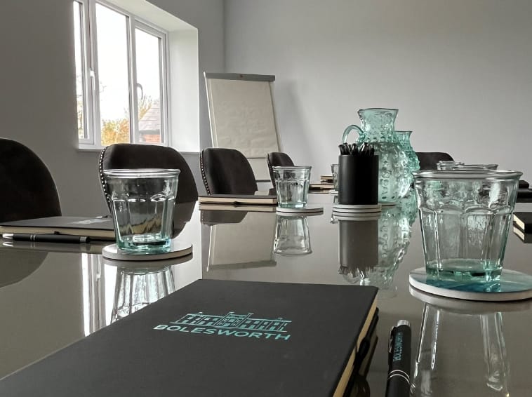 A boardroom desk with pens, notepads, drinking glasses, jug of water sand flip chart