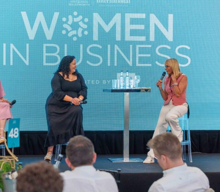 Three women seated on a stage engage in a discussion during a Women in Business panel, with a Bolesworth backdrop in view.