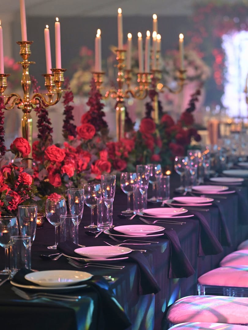 A beautifully arranged candlelit wedding reception table, featuring gold candelabras, rich floral arrangements with deep red roses, elegant table settings with black napkins, and crystal-clear glassware, creating a romantic and luxurious ambiance.