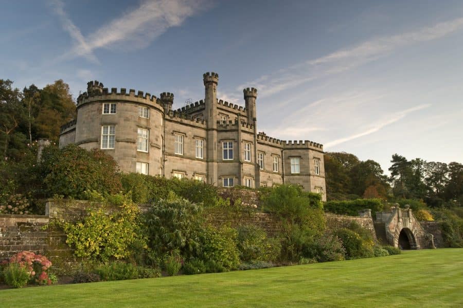 A stunning view of Bolesworth Castle, a historic estate in Cheshire, UK, surrounded by lush gardens and an expansive lawn. The castle's majestic stone architecture and picturesque landscape create an idyllic setting.