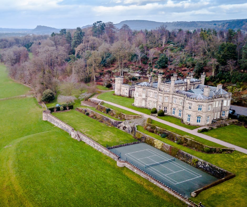 A large Castle with turrets surrounded by trees and gardens and a tennis court