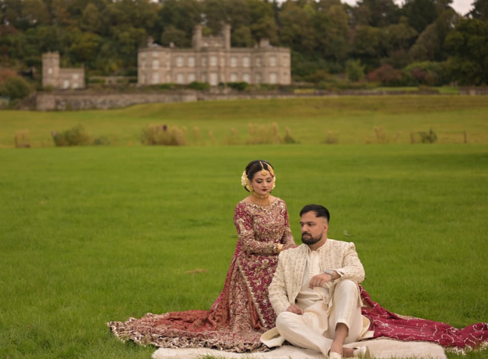 Bride and groom in traditional South Asian wedding attire, seated on a picnic blanket with Bolesworth Castle in the background.